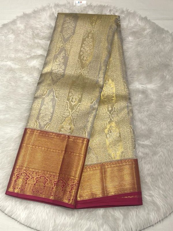 A brightly lit, folded traditional Indian silk saree featuring a cream or light silver-grey body with vertical panels of intricate gold and silver-gold zari brocade weaving, possibly with paisley or ornamental motifs. The saree has a wide, contrasting border in a deep copper or orange-gold tone with horizontal gold zari stripes and an inner band of woven traditional motifs, finished with a narrow, solid magenta or dark pink piping along the edge.