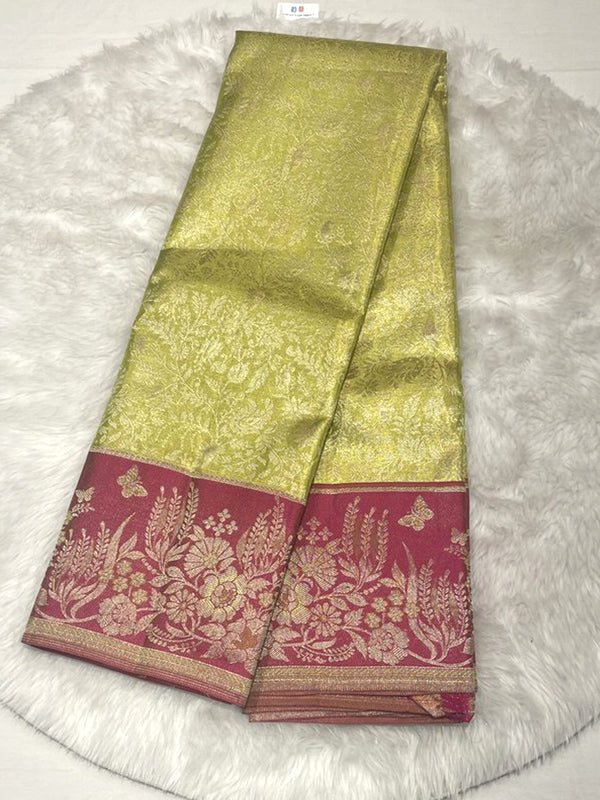A brightly lit, folded traditional Indian silk saree featuring a shimmering lime-green or chartreuse body with all-over intricate gold zari floral and leaf brocade weaving. The saree has a wide, contrasting border in deep magenta or hot pink, which is densely woven with large gold zari floral, vine, and butterfly motifs. The saree is displayed on a fluffy white or off-white textured rug.
