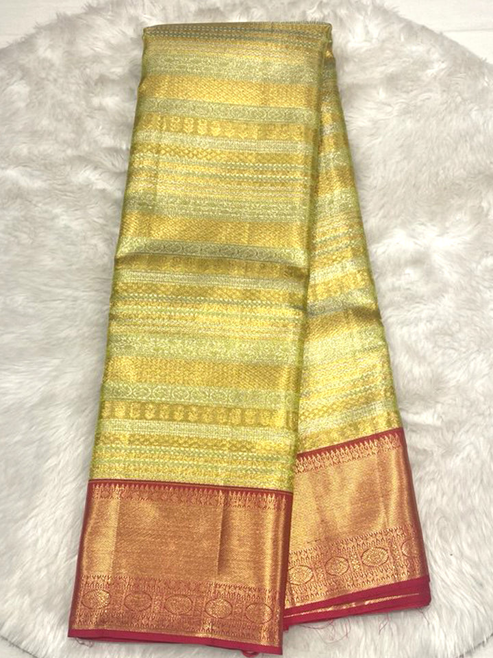 A brightly lit, folded traditional Indian silk saree featuring a shimmering lime-green and golden striped body with intricate brocade weaving. The saree has a wide, contrasting deep gold/copper border with fine woven motifs, finished with a narrow, solid magenta or dark pink piping along the edges. The saree is displayed on a fluffy white or off-white textured rug.
